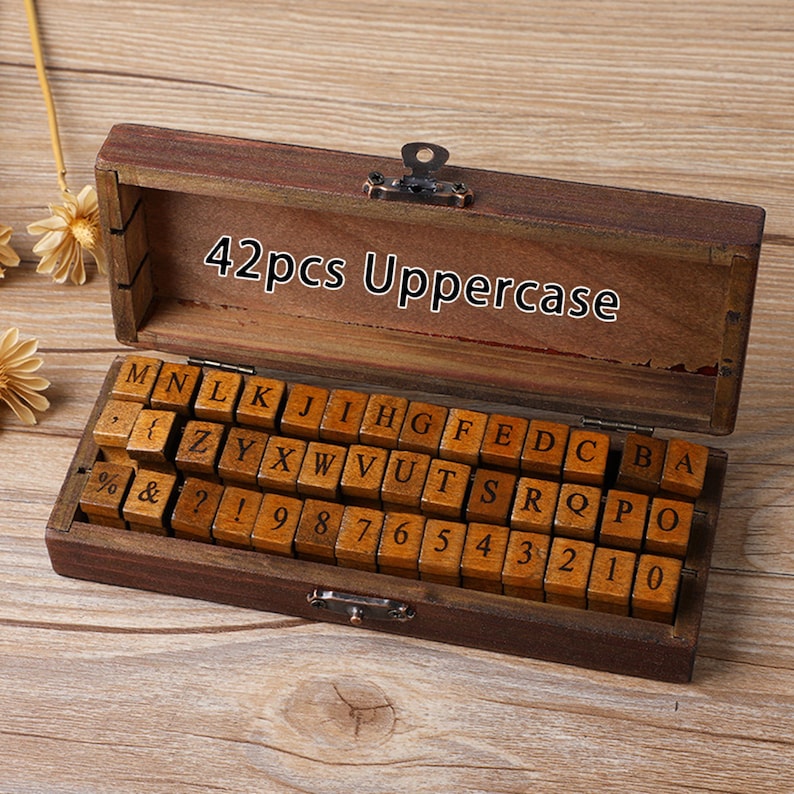 May include: A wooden box containing 42 uppercase letter stamps and numbers. The stamps are arranged in rows inside the open box. The box is made of dark brown wood and has a metal clasp. The text "42pcs Uppercase" is printed on the inside of the lid.