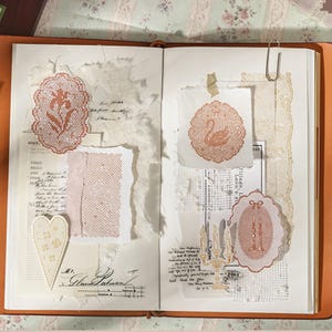May include: An open journal featuring collage elements including orange lace-style die-cut shapes of a flower, a swan, and candles. The pages contain torn white paper, handwritten script, grid patterns, a heart-shaped cutout, and a silver paperclip.