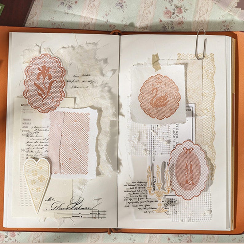 May include: An open journal featuring collage elements including orange lace-style die-cut shapes of a flower, a swan, and candles. The pages contain torn white paper, handwritten script, grid patterns, a heart-shaped cutout, and a silver paperclip.