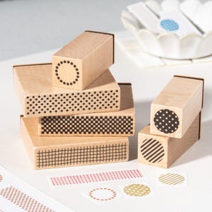 May include: A collection of wooden stamps with various geometric patterns, including dots, grids, and stripes. The stamps are light brown with black ink designs. Small sticker labels with matching patterns are also present.