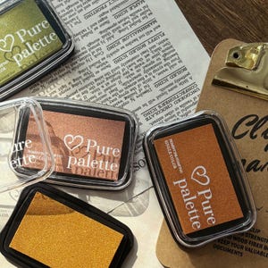 May include: Close-up of several rectangular pigment ink stamp pads in clear plastic cases. The pads are in various colors, including gold, copper, and olive green. Each pad has the text "Pure palette" printed on it.