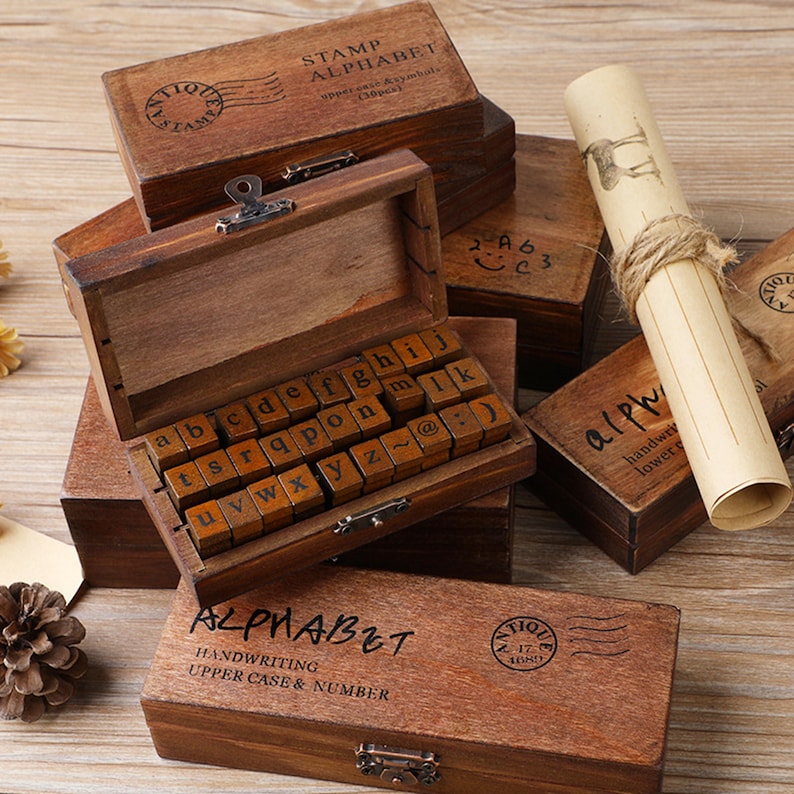 May include: Wooden alphabet stamp sets with uppercase and lowercase letters, numbers, and symbols. The stamps are stored in small, antique-style wooden boxes. One box is open, revealing the letter stamps. The boxes are brown with black text.