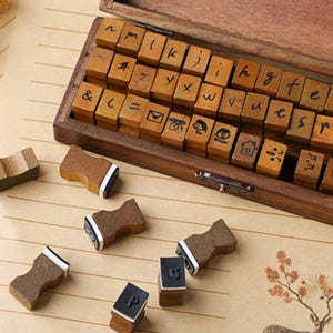 May include: A wooden alphabet stamp set with individual letter and symbol stamps. The stamps are arranged in a wooden box, with some stamps scattered around. The stamps have a dark brown wooden handle and a white and black stamp pad.