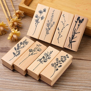 Peut inclure: Ensemble de six tampons en bois avec des motifs botaniques. Chaque tampon a un bloc de bois rectangulaire avec une illustration florale noire. Les tampons sont disposés sur une surface en bois, avec des fleurs séchées et des bâtons de bambou en arrière-plan.