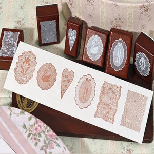 May include: A collection of six rectangular wooden stamps with dark brown handles, each featuring a unique white lace pattern on the top. A white paper strip displays six corresponding copper-colored ink impressions of the lace designs.