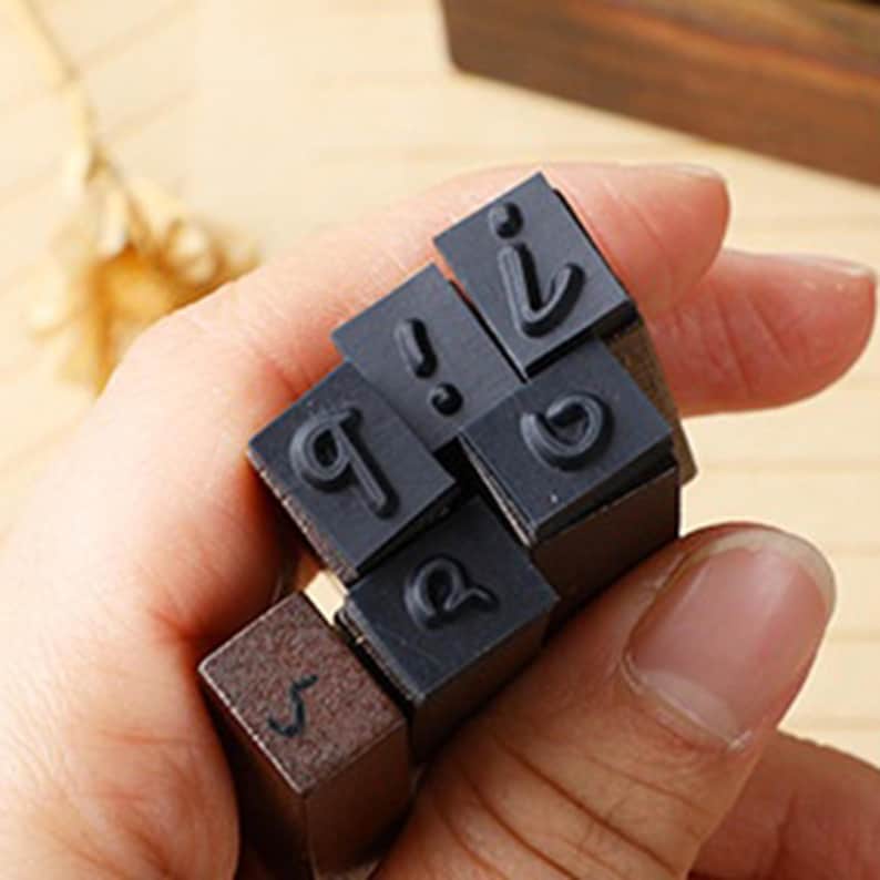 May include: A collection of small, square letter stamps with black rubber letter faces and wooden blocks. The stamps include the letters "s", "q", "b", and punctuation marks. The stamps are held in a hand.