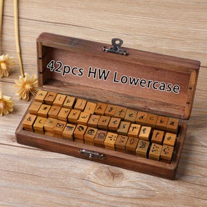 May include: A wooden box containing 42 lowercase letter stamps. The stamps are arranged in rows inside the open box. The box is made of dark brown wood and has a metal clasp. The text "42pcs HW Lowercase" is printed on the lid.