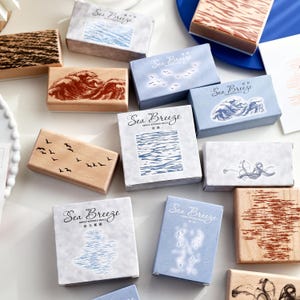 Puede incluir: Un conjunto de sellos de madera y cajas azules con el texto "Sea Breeze". Los sellos presentan varios diseños, incluyendo olas, pájaros y patrones abstractos. Las cajas son de color azul claro y tienen el texto "Sea Breeze".