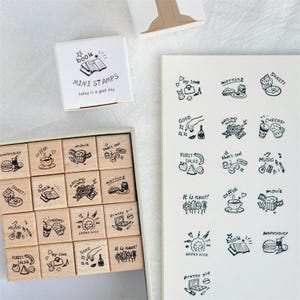 May include: A collection of wooden mini stamps featuring hand-drawn illustrations and phrases related to everyday life, such as food, drinks, and activities. The set includes a box of stamps and a pad with stamped images. The box reads "MINI STAMPS today is a good day."