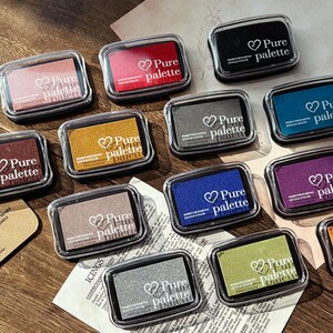 May include: A collection of rectangular ink pads in various colors, including red, black, and gold, each encased in a clear plastic container. The text "Pure palette" is printed on each pad, along with a heart symbol. The pads are arranged on a wooden surface.