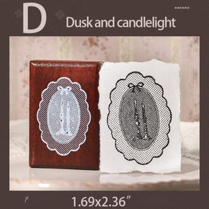 May include: A rectangular wooden stamp with a dark brown finish featuring a scalloped oval design of two candles. Next to the stamp is a white paper impression showing the same black ink design. The stamp measures 1.69 by 2.36 inches.
