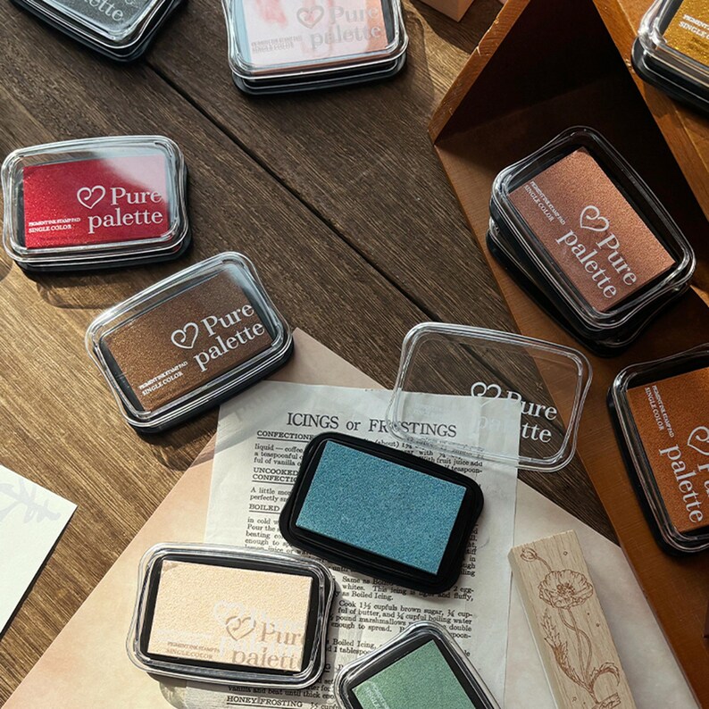 May include: A collection of rectangular ink pads in various colors, including red, brown, and teal, with clear plastic lids. Each pad is labeled "Pure palette" with a heart symbol. The pads are arranged on a wooden surface, suggesting a craft or stationery theme.