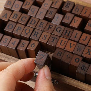 May include: Wooden alphabet and number stamps in a wooden box. The stamps are dark brown with black lettering. A hand is holding a stamp with the number 7. The set includes letters, numbers, and symbols for crafting and scrapbooking.