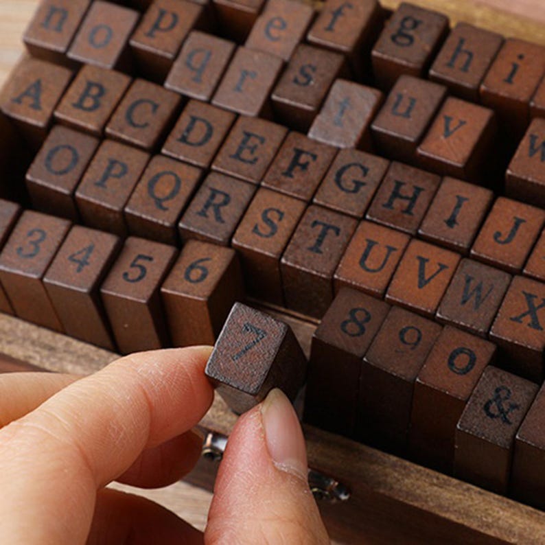May include: Wooden alphabet and number stamps in a wooden box. The stamps are dark brown with black lettering. A hand is holding a stamp with the number 7. The set includes letters, numbers, and symbols for crafting and scrapbooking.