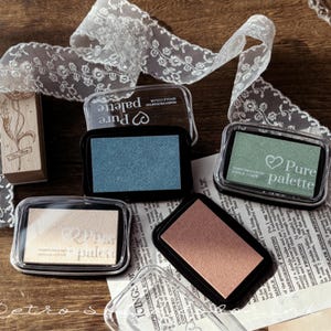 May include: A collection of Pure Palette ink pads in various colors, including blue, green, and peach, with clear plastic lids. A decorative lace ribbon and a wooden stamp are also present, suggesting a crafting theme.