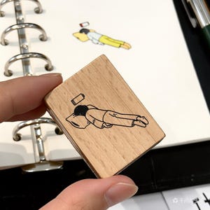 Peut inclure: Tampon en bois avec une illustration noire d'une personne allongée sur un oreiller, avec un symbole de batterie faible au-dessus de la tête. Le tampon est tenu dans une main, avec un carnet et un stylo en arrière-plan.