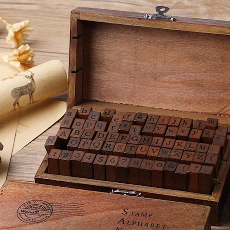 May include: A wooden stamp set with uppercase and lowercase letters, numbers, and symbols. The stamps are arranged in a wooden box with a hinged lid. The box also contains a stamp pad. The text "STAMP ALPHABET" is visible on the box.
