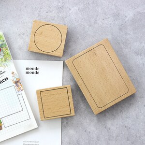 Könnte beinhalten: Holzstempel mit schwarzen Umrissen eines Kreises, Quadrats und Rechtecks. Die Stempel sind auf einer weißen Oberfläche mit dem Text "monde monde" und einem Rastermuster angeordnet. Die Stempel sind aus hellem Holz gefertigt.