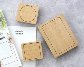 May include: Wooden stamps with black outlines of a circle, square, and rectangle. The stamps are arranged on a white surface with the text "monde monde" and a grid pattern. The stamps are made of light-colored wood.