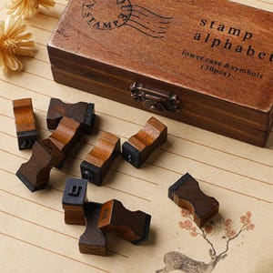 May include: A wooden box set of alphabet stamps with the text "stamp alphabet lower case & symbols (30pcs)". The stamps are made of wood and have black rubber stamp pads. The box is brown with a metal clasp.