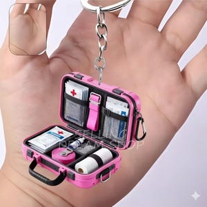 May include: A pink miniature first aid kit keychain, open to reveal various medical supplies. The kit includes bandages, tape, and other essentials. It has a black handle and a silver keychain ring.