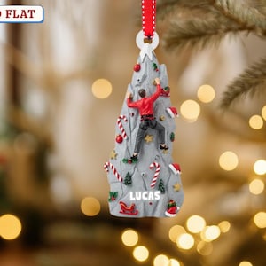 May include: A gray Christmas ornament shaped like a mountain with a climber in a red jacket. Candy canes, Santa hats, and a sled decorate the mountain. The name "LUCAS" is printed on the ornament. A red ribbon is attached for hanging.