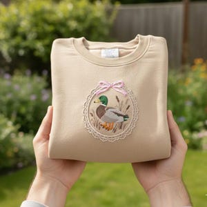 May include: A tan sweatshirt with an embroidered design of a mallard duck inside a decorative oval frame with a pink bow. The duck has green, brown, and gray feathers. The sweatshirt is folded and held by hands.