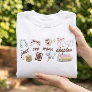 May include: White sweatshirt with embroidered design. The design includes headphones, glasses, a coffee mug, a bow, a candle, a tablet, flowers, and books. The text "just one more chapter" is also embroidered on the sweatshirt.