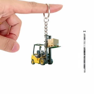 May include: A yellow and black forklift keychain with a silver chain and ring. The forklift is carrying a small cardboard box on a pallet. The keychain is held up by a finger.