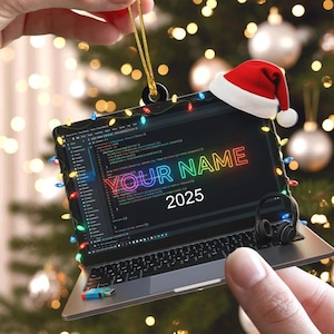 May include: A laptop-shaped Christmas ornament with a festive design. The screen displays code and the text "YOUR NAME" in neon colors, with "2025" below. A small Santa hat sits on top, and the laptop is adorned with colorful lights.