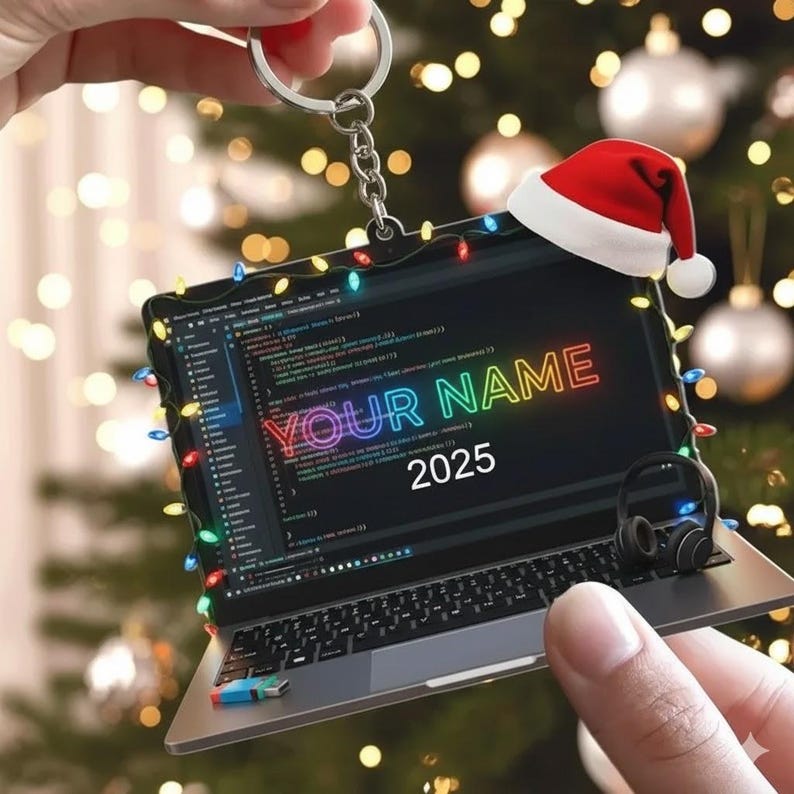 May include: Laptop-shaped Christmas ornament with a Santa hat, string lights, and the text "YOUR NAME 2025." The laptop screen displays code. A small pair of headphones is on the side. The ornament is attached to a keychain.