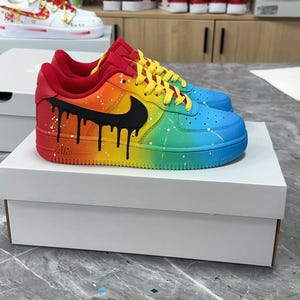 Pode incluir: T&ecirc;nis personalizados com arco-&iacute;ris, com um logotipo preto e um design de gotejamento preto. Os sapatos t&ecirc;m detalhes em vermelho, amarelo e azul. Os t&ecirc;nis s&atilde;o exibidos em uma caixa branca.