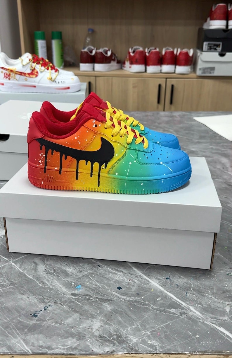 Pode incluir: T&ecirc;nis personalizados com arco-&iacute;ris, com um logotipo preto e um design de gotejamento preto. Os sapatos t&ecirc;m detalhes em vermelho, amarelo e azul. Os t&ecirc;nis s&atilde;o exibidos em uma caixa branca.