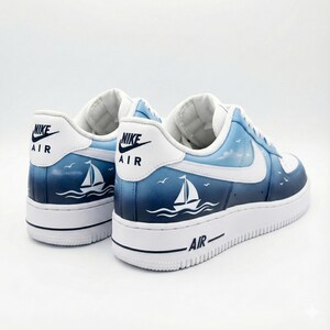 Puede incluir: Un par de zapatillas blancas y azules con tem&aacute;tica n&aacute;utica. Los zapatos presentan un dise&ntilde;o azul degradado con un velero y p&aacute;jaros. El logotipo de Nike es visible en el tal&oacute;n.