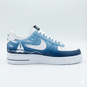 Puede incluir: Una zapatilla Air Force 1 blanca y azul con un dise&ntilde;o n&aacute;utico. La zapatilla presenta un logotipo de Nike blanco, un velero y una escena de cielo y oc&eacute;ano azul degradado. La suela es blanca.