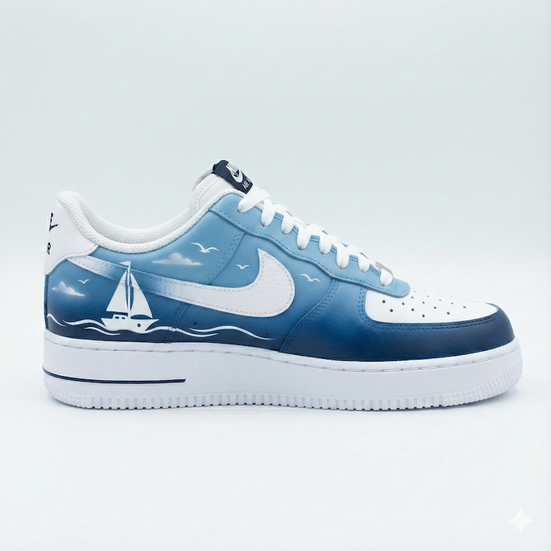 Puede incluir: Una zapatilla Air Force 1 blanca y azul con un dise&ntilde;o n&aacute;utico. La zapatilla presenta un logotipo de Nike blanco, un velero y una escena de cielo y oc&eacute;ano azul degradado. La suela es blanca.