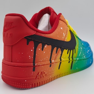 Pode incluir: Um t&ecirc;nis Nike Air Force 1 pintado sob medida. O t&ecirc;nis apresenta um gradiente de arco-&iacute;ris vibrante, passando do vermelho ao azul, com detalhes de gotejamento preto e respingos brancos. O logotipo da Nike &eacute; vis&iacute;vel em preto.
