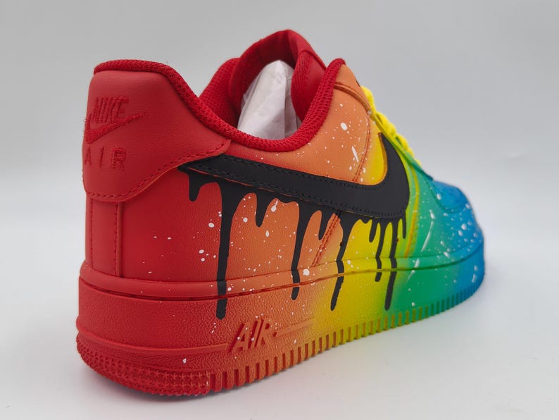 Pode incluir: Um t&ecirc;nis Nike Air Force 1 pintado sob medida. O t&ecirc;nis apresenta um gradiente de arco-&iacute;ris vibrante, passando do vermelho ao azul, com detalhes de gotejamento preto e respingos brancos. O logotipo da Nike &eacute; vis&iacute;vel em preto.