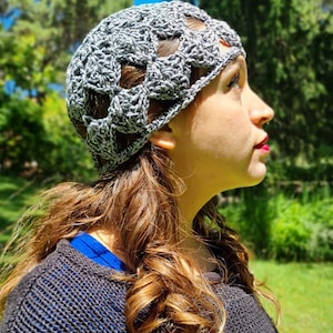 May include: A gray crocheted hat with an open weave design. The hat is worn by a person with long, wavy brown hair. The person is wearing a dark gray sweater with a blue shirt underneath. The background is a sunny outdoor scene with green trees.
