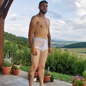 Peut inclure: Sous-vêtements blancs en résille avec une ceinture bleue et blanche. Les sous-vêtements sont faits d'un matériau transparent. La personne se tient sur un patio carrelé avec des plantes en pot et un arrière-plan panoramique.