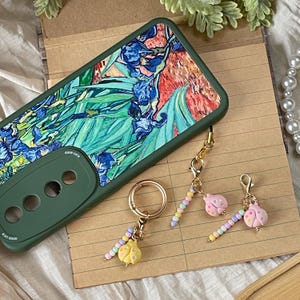 Könnte beinhalten: Eine grüne Handyhülle mit floralem Gemäldedesign, ein goldener Schlüsselanhänger mit gelbem Marienkäfer-Anhänger und zwei Marienkäfer-Anhängern mit Perlenakzenten. Auf der Handyhülle steht "Good Luck".
