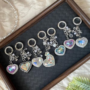 Pode incluir: Seis chaveiros em forma de coração com cristais iridescentes e detalhes em metal prateado. Cada chaveiro apresenta um pingente grande em forma de coração brilhante com uma borda transparente cravejada de pedras preciosas. Os chaveiros estão dispostos sobre uma superfície escura e texturizada.