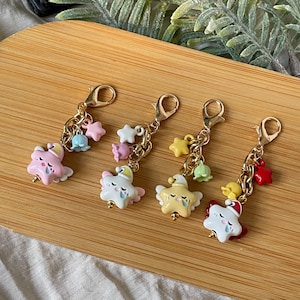 Può includere: Quattro ciondoli a forma di stella con catene e chiusure color oro. Ogni ciondolo presenta una diversa combinazione di colori, tra cui rosa, bianco, giallo e rosso, con piccoli accenti a forma di stella e campana. I ciondoli sono su una superficie di legno.