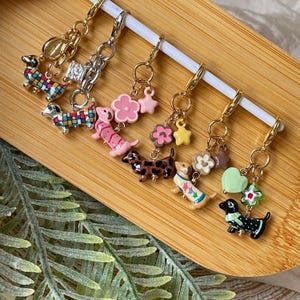 Può includere: Una collezione di orecchini con ciondoli a forma di bassotto colorati. Ogni orecchino presenta un bassotto in miniatura con vari motivi e colori, abbinato a ciondoli a forma di fiori e stelle. Gli orecchini sono esposti su una superficie di legno.