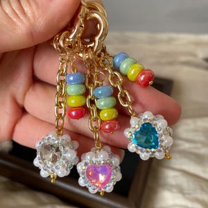 Peut inclure: Porte-clés doré avec plusieurs breloques. Les breloques comprennent des perles empilées colorées et des pendentifs en cristal en forme de cœur et ronds, le tout orné de fausses perles. Le porte-clés est tenu par une main.