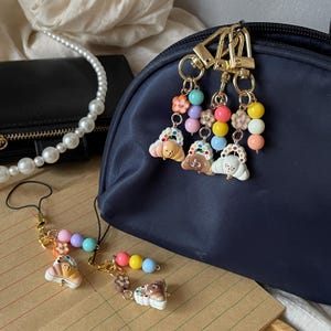 May include: A selection of colourful keychains and charms featuring small dog-shaped pendants with beaded accents. The keychains are attached to a navy blue bag and a gold-coloured clasp. Additional charms with colourful beads and flower accents are also present.