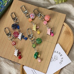 May include: A collection of decorative keychains with silver-tone clasps. Each keychain features a charm with colorful beads, flowers, umbrellas, and stars. The keychains are arranged on a brown surface.