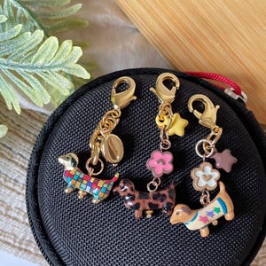 Könnte beinhalten: Drei Dackel-Anhänger mit goldfarbenen Verschlüssen. Ein Anhänger hat ein buntes Karomuster, ein anderer ein braunes und schwarzes Muster und der dritte ein Blumenmuster. Jeder Anhänger ist mit einem goldfarbenen Verschluss mit zusätzlichen Anhängern versehen.