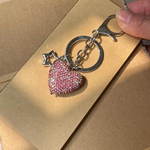 May include: A silver keychain with a pink rhinestone heart pendant and a small star charm. The heart-shaped pendant is covered in small, sparkling pink rhinestones. The keychain is attached to a silver clasp and chain.