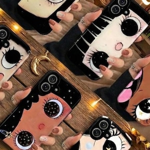 May include: A collection of black phone cases with unique designs. Each case features a cartoon-style face with large eyes, eyelashes, and a crescent moon. The cases have a black border and a variety of skin tones and hair colors.